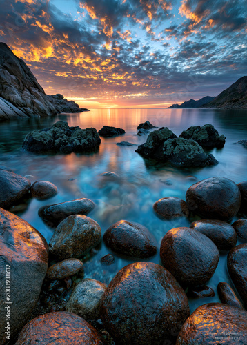 Waves gently lap at smooth stones along the shoreline as the sun sets, painting the sky with hues of orange and purple. Clouds reflect on calm waters, creating a peaceful scene.