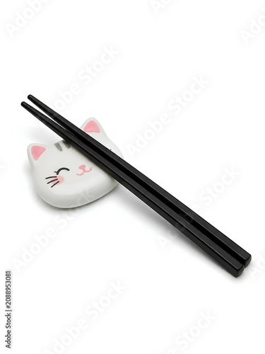 A pair of sleek black chopsticks rest on a ceramic chopstick holder shaped like a cat's face. The holder features a cute, smiling cat design with pink ears and closed eyes, adding a playful element.