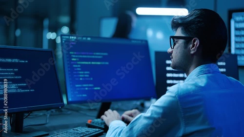 Focused software developer in glasses coding on multiple monitors in a dark high-tech office. Male IT professional analyzing programming data late at night. Cyber security expert working fast.