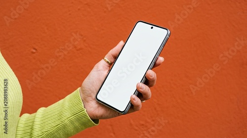 Close up of a hand wearing a ring holding a modern smartphone with a blank white screen against a textured orange wall with communication touchscreen technology background electronic minimalist