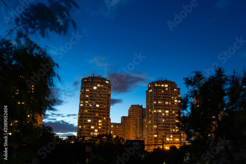Urban district. High residential buildings in front of a dark blue evening summer sky with tree silhouettes. City at night