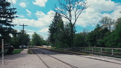 Train tracks on a rocky road surrounded by green trees, a blue sky, power lines, and flying birds.