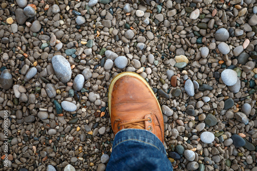 Quadro su tela First Person View of a Boot Standing on a Pebble Beach