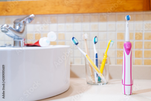 Photography Electric toothbrush standing on a bathroom counter next to a sink with a tube of toothpaste and a glass holding several manual toothbrushes