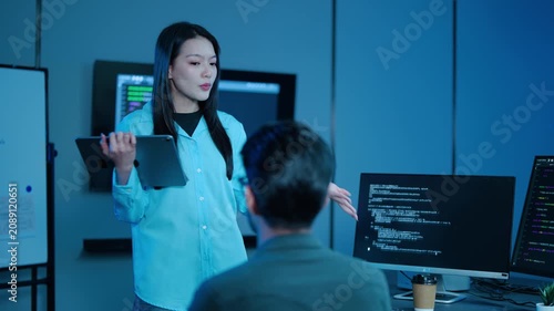 Asian female IT project manager holding a tablet discusses software coding on computer monitors with a male developer working late in a modern dark blue tech office room. Teamwork collaboration.