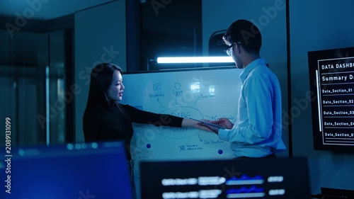 Two Asian programmers brainstorming software architecture on a whiteboard in a dark high-tech office. Tech team discussing coding flow late at night. IT developers planning application logic.