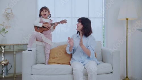 A mother sits on a couch with her young daughter. The girl plays a ukulele while her mom looks at her and shares a smile. They enjoy a happy moment together in their home.