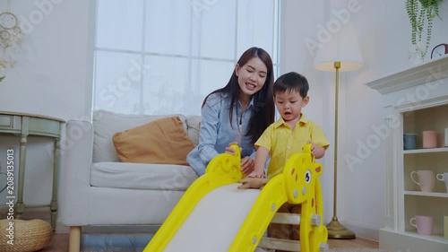 A mother sits beside her young son as he plays on a small slide. They are in a bright living room with a sofa and plants. They share smiles and laughter together.