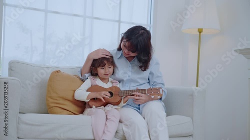 A mother sits on a couch with her young daughter. The girl plays a ukulele while her mom looks at her and shares a smile. They enjoy a happy moment together in their home.