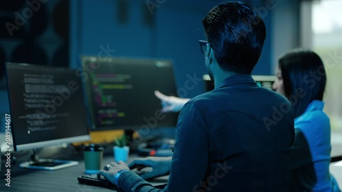 Two asian expert software developers collaborating on complex code at night. Man types and points at multiple monitors while a woman observes in a dark modern office. IT team working late on a project