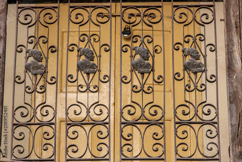An ornate wrought iron window grill featuring decorative scrollwork and metallic profile busts of Chhatrapati Shivaji Maharaj, set against a classic yellow paneled wooden door with a padlock.