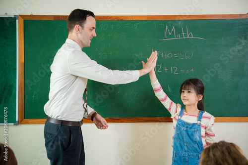 Friendly male teacher giving high five to a cute Asian schoolgirl in front of a chalkboard. Happy schoolteacher celebrating success with a primary student after solving a math problem.
