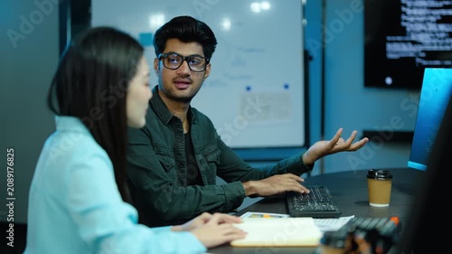 Diverse software developers discuss coding projects while working late at night in a modern tech startup office. Asian male programmer types on his computer keyboard as the female colleague helps.