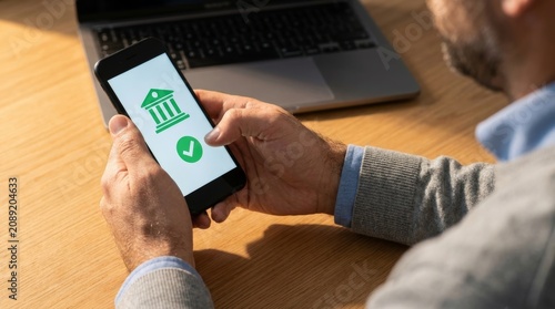 a mobile banking app on a smartphone to confirm a transaction at a wooden desk with a laptop.