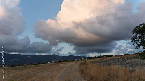 Large storm cloud hanging over a dry rural road, hay bales and distant mountains.