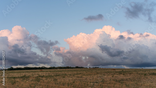 Dry rural field under pastel sunset clouds and a dramatic rain front on the horizon.