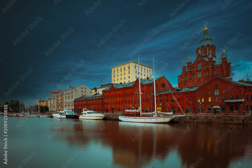 Obraz premium dramatic view of the historic red brick Uspenski Cathedral and surrounding buildings reflected in the calm waters of the harbor under a spectacular dark sunset sky.
