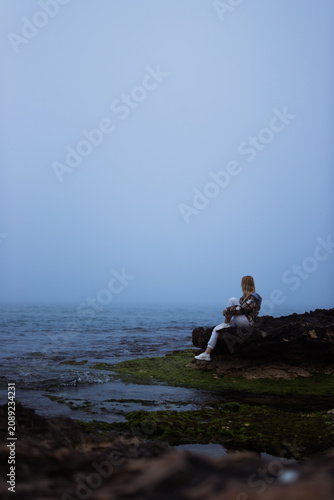 Fotografie Young woman sitting calmly on coastal rocks watching the quiet sea disappear int