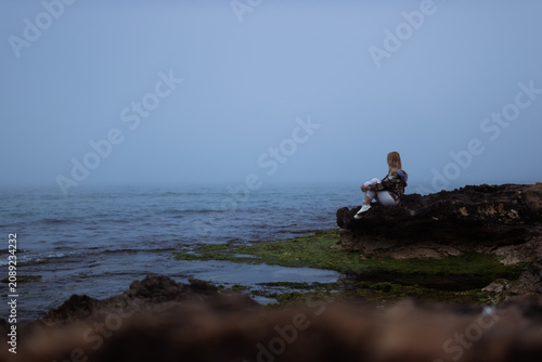 Принти на полотні Young woman sitting on coastal rocks, contemplating ocean horizon on a misty day