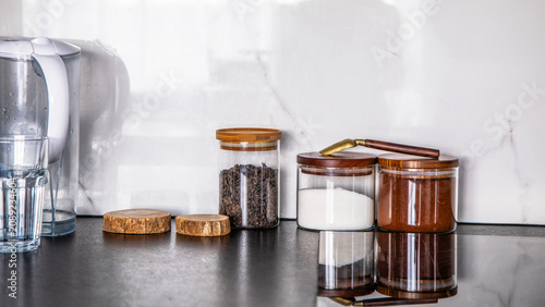 Schilderij op canvas Glass canisters with acacia lids storing tea, sugar, and coffee on a dark kitchen counter with a water filter jug and eco-friendly coasters