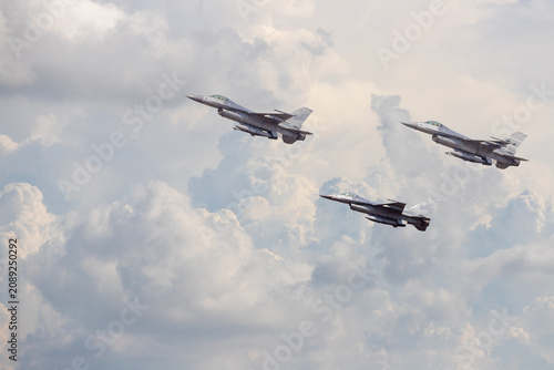 Konstfotografi Three military fighter jets fly in formation against a bright blue sky with large puffy white clouds
