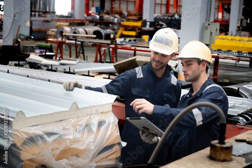 Two factory workers in hard hat and coverall inspecting metal roofing panels on production line, discussing quality control and using tablet to record measurements and findings