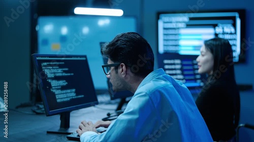 Focused young asian male IT software engineer with glasses typing code on computer monitor in a high-tech dark control room with blue light, while a professional female colleague works in the office 