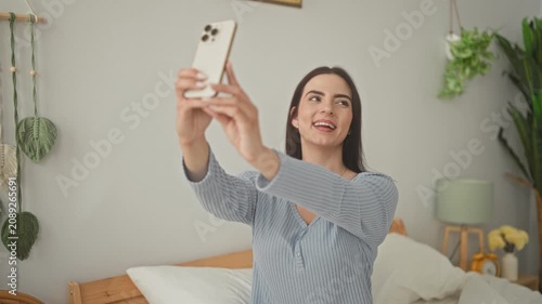 Woman holding smartphone and extending arms for a selfie, puckered lips and smiling expression on bed in a bedroom; playful confidence.