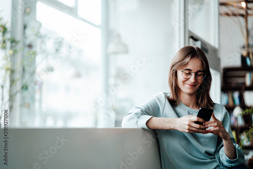Smiling woman text messaging through smart phone sitting in cafe