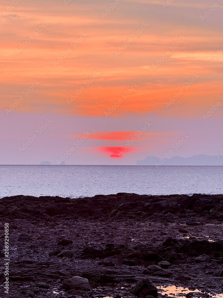 Obraz premium Stunning tropical sunset with a glowing red sun sinking below the horizon, soft pastel clouds, calm ocean, and rocky shoreline creating a peaceful coastal landscape.