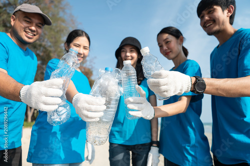 Group of happy Asian volunteers holding trash bags and plastic bottles on the beach, smiling at camera during a successful community cleanup event.