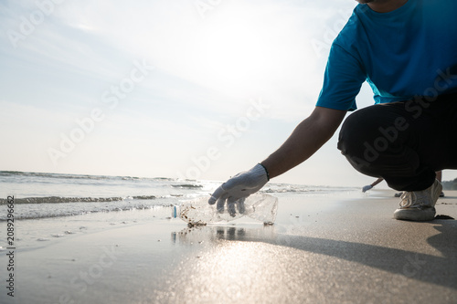 Close-up of a volunteer's hand wearing a glove picking up a discarded plastic bottle from the sea shore, highlighting ocean pollution and environmental cleanup efforts.