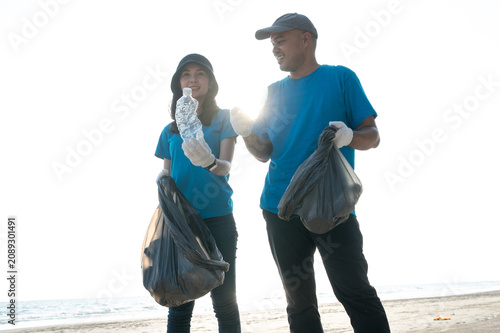 Two happy volunteers smiling and talking while collecting plastic bottles into a trash bag on the beach, enjoying their environmental community service together.