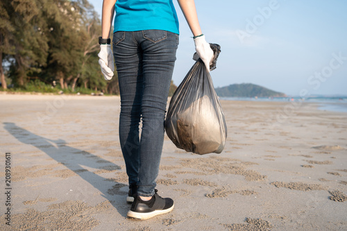 Young female volunteer collecting plastic bottles into a trash bag on the beach, backlit by bright sunlight, promoting marine conservation and eco friendly lifestyle.