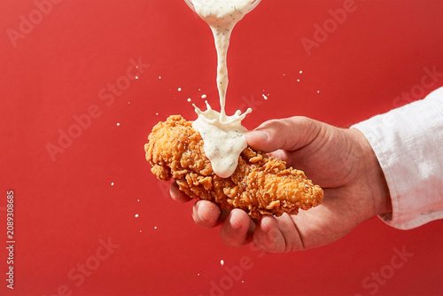 Obraz na płótnie Hand Holding Crispy Chicken Tenders with Ranch Sauce Pouring on Red Background A