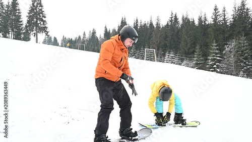Two snowboarders prepare on a snow-covered slope. One assists the other. Trees frame the background in a winter wonderland setting.