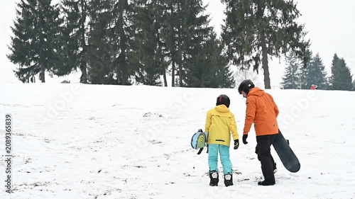 A snowboard instructor patiently assists a child on a snowy hillside. Other winter sports enthusiasts are visible in the background, enjoying the snowy landscape.