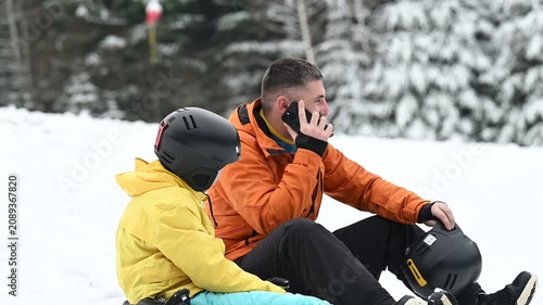A man speaks on his phone while sitting beside a child wearing a ski helmet in the snow. They appear relaxed and happy, enjoying a winter day outdoors.