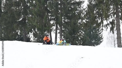 Two individuals, one appearing male and the other female, take a break from snowboarding. They are seated in the snow at the bottom of a hill, with a backdrop of tall evergreens.