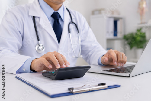 Close up of a medical professional calculating expenses. Doctor hands using a calculator and laptop for patient insurance claims, medical bills, and clinic budget planning.