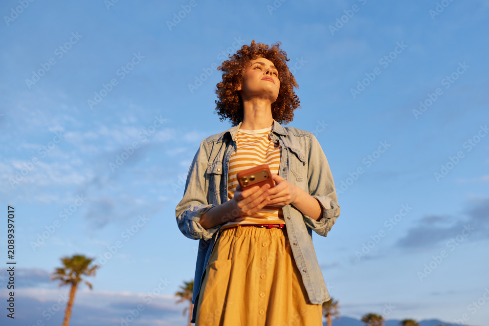 Fototapeta premium A confident woman stands outdoors in bright daylight, looking up while holding a phone. A denim jacket and striped top complete a casual lifestyle portrait.