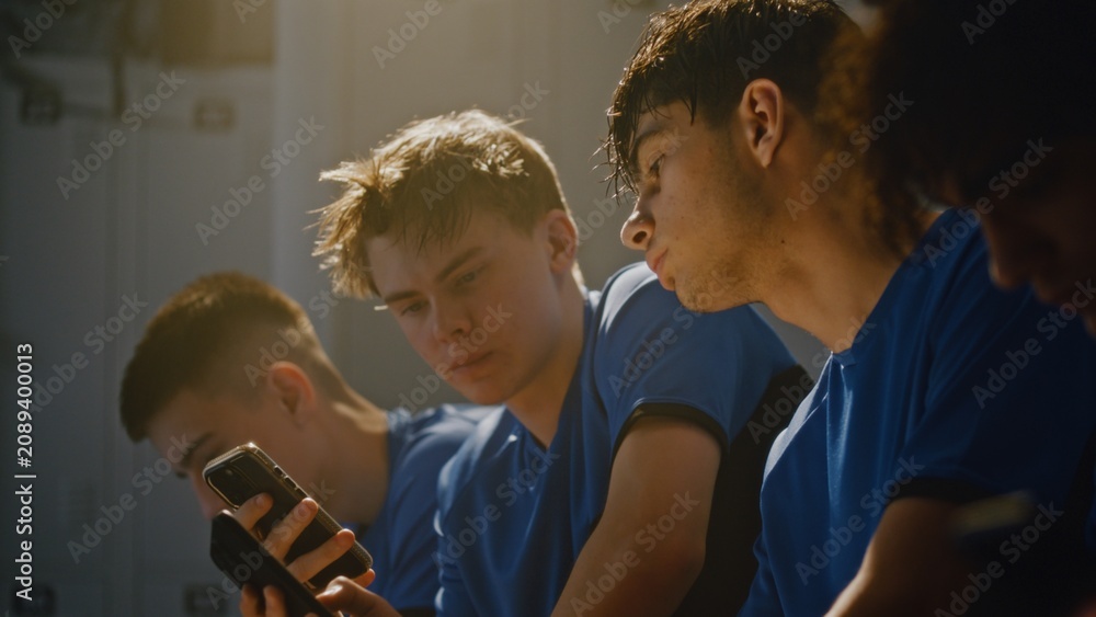Fototapeta premium Young Male Athletes in Soccer Kits Gathered Together on Bench in Locker Room, Looking Mobile Phone and Discussing Something With Smiles. Concept of Youth Connectivity, Team Unity, and Digital Sharing.