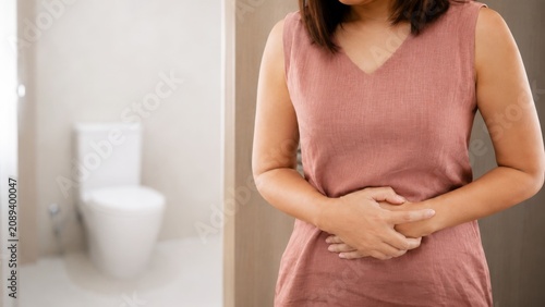 A woman in a pink shirt clutches her stomach, looking uncomfortable, with a bathroom visible in the background.