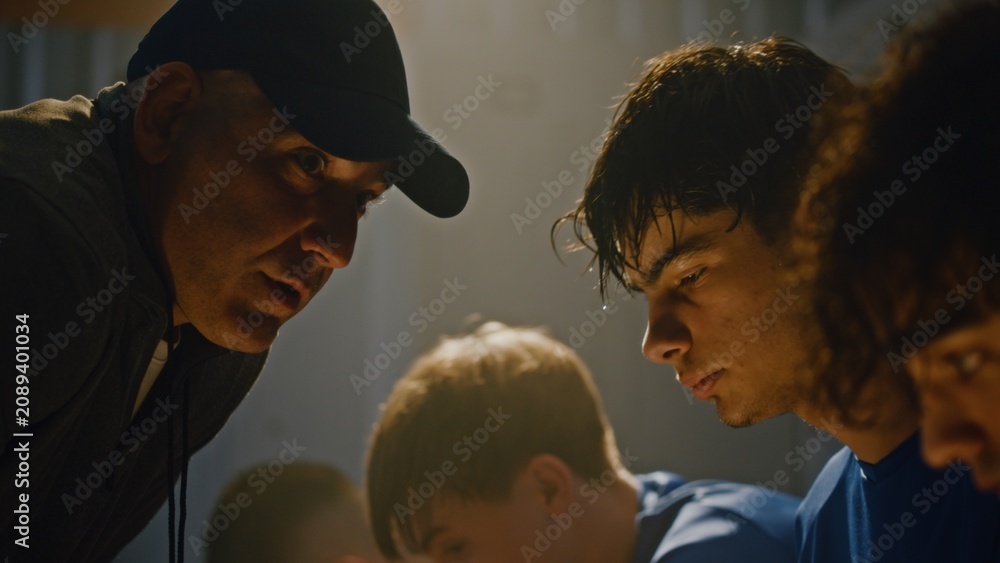 Fototapeta premium Strict Football Manager in Cap Aggressively Lecturing Team in Row of Lockers. Stands Over Seated Teenagers to Demand Better Performance. Crisis Moment Within Sports Team. Leadership Under Pressure.