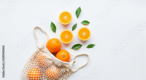 Fresh oranges in reusable cotton mesh bag, organic citrus fruit halves with green leaves, zero waste grocery shopping concept on white background