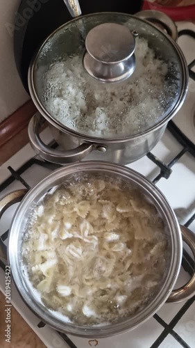 Boiling farfalle pasta and steaming white rice cook in stainless steel pots on a gas stovetop. Concept of homemade family meals, domestic culinary routines, and everyday multitasking.