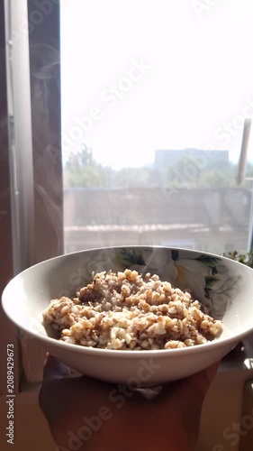 A person holds a white bowl of hot buckwheat porridge with visible steam in front of a bright window. Concept of healthy lifestyle, nutritious morning meal, and balanced breakfast traditions