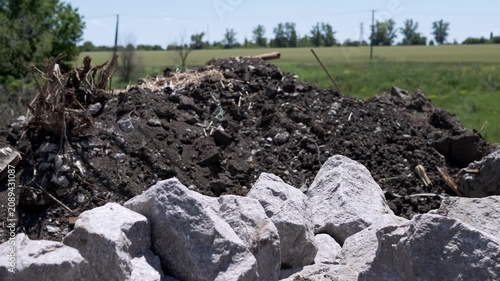 Grey rocks sit before a pile of dark excavated soil and debris in a sunny green field. Concept of construction preparation, environmental impact, and industrial landscaping works.