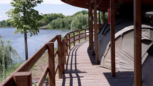 A row of modern camping tents stands under a protective wooden pavilion on a boardwalk next to a calm lake. Concept of luxury camping, summer vacation, and peaceful outdoor recreation.