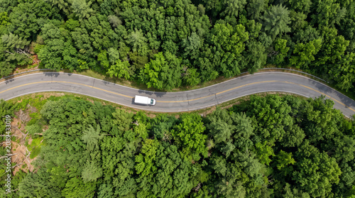 Aerial view of a white delivery truck driving on a winding asphalt road through a dense green forest during summer